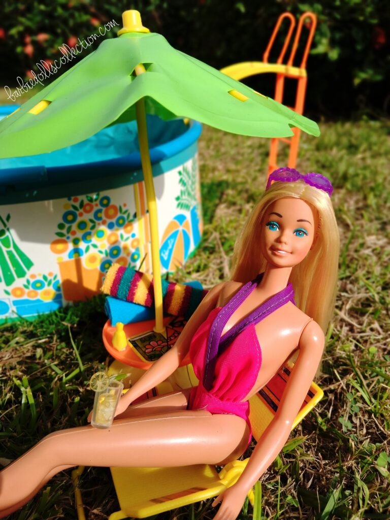 Malibu Barbie relaxing poolside in her swimsuit while sitting under a patio umbrella, holding a lemonade with beach towels and sunscreen nearby.